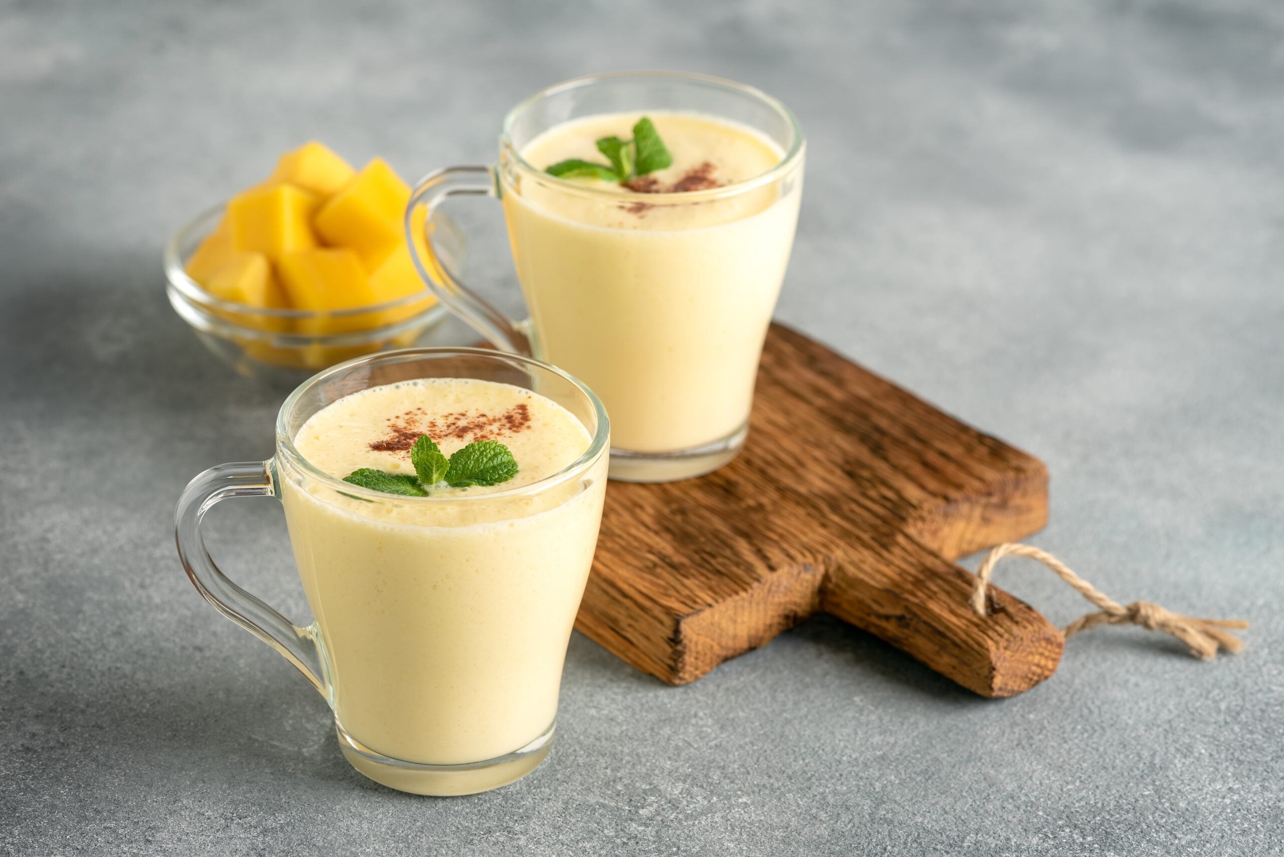A concrete table showcasing glasses of mango Lassi and some fresh mango. 