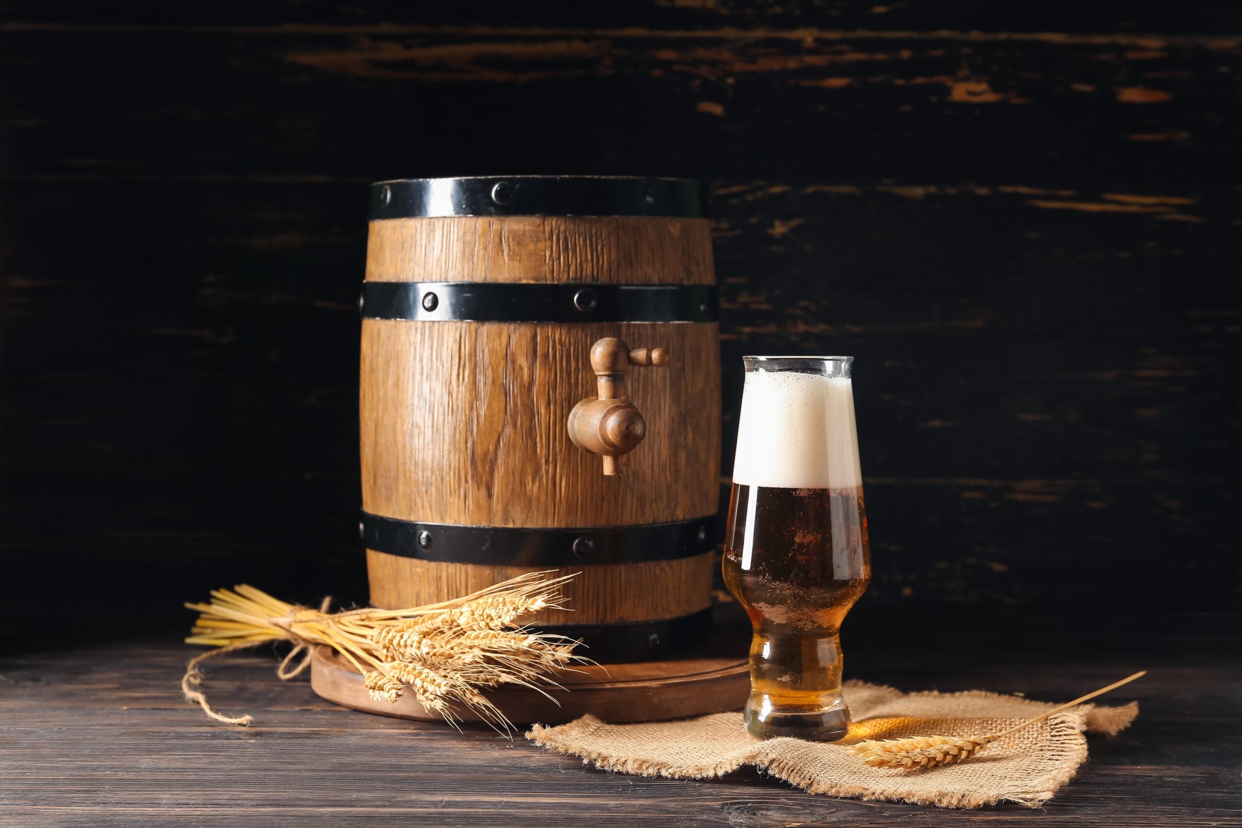 Wooden barrel with tap, beer glass, wheat stalks on burlap.