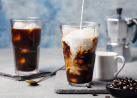 Two glasses of iced coffee, one with cream being poured, moka pot, coffee beans, and spoon.