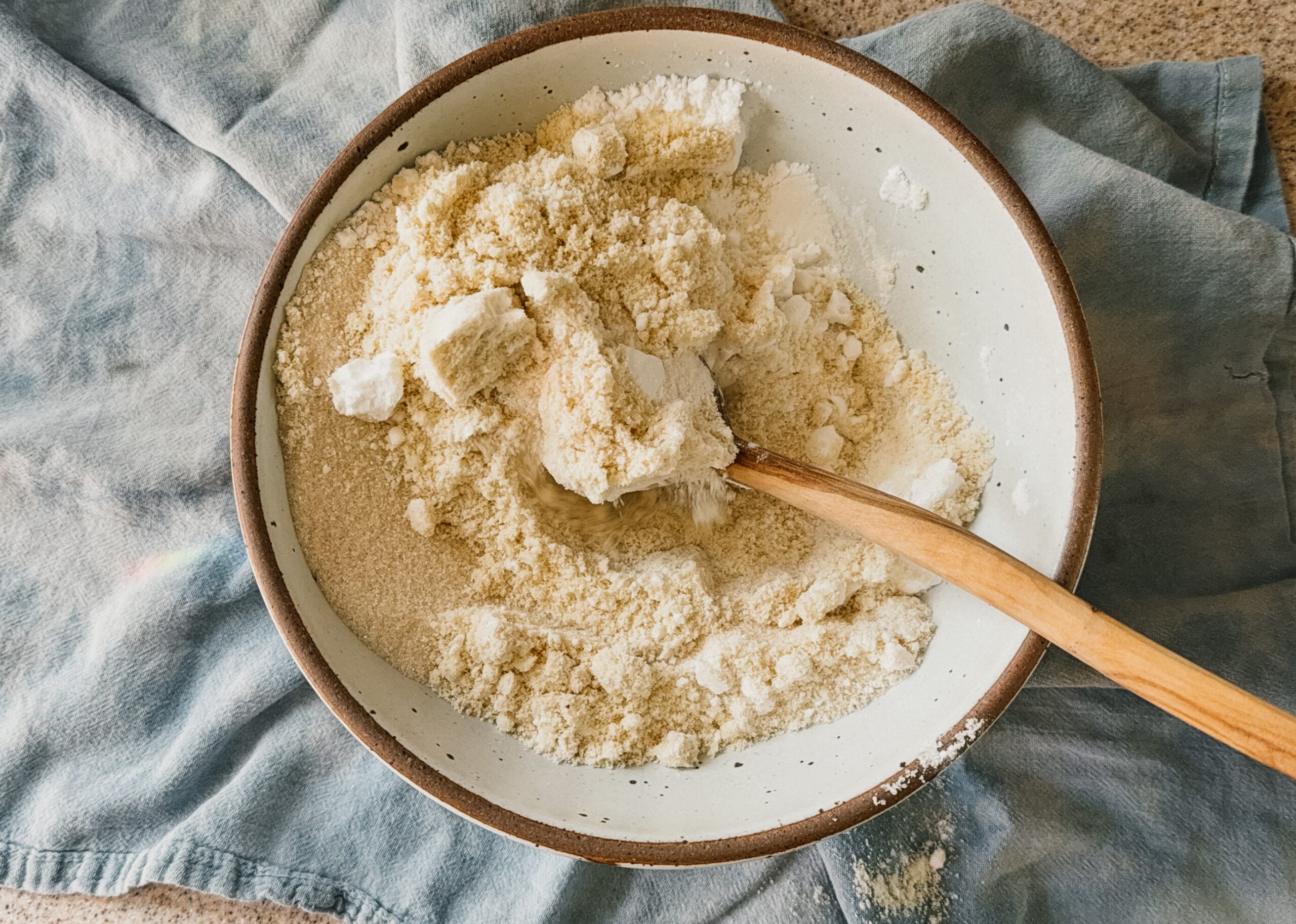 A bowl of ingredients being mixed with a wooden spoon. A blue tea towel lays underneath the bowl.
