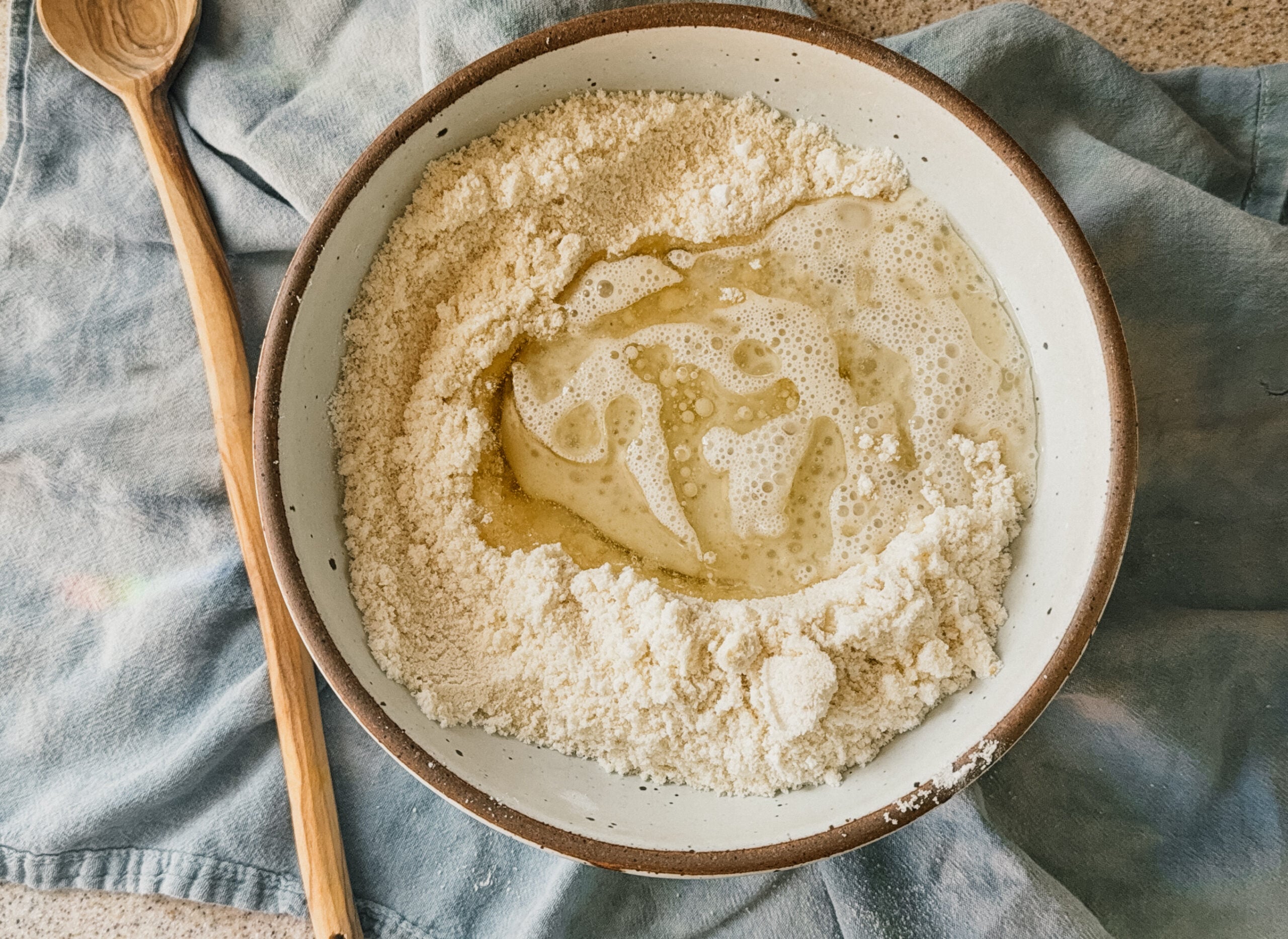 A bowl of ingredients about to be mixed. A wooden spoon rests to the left of the bowl and a blue tea towel lays underneath.