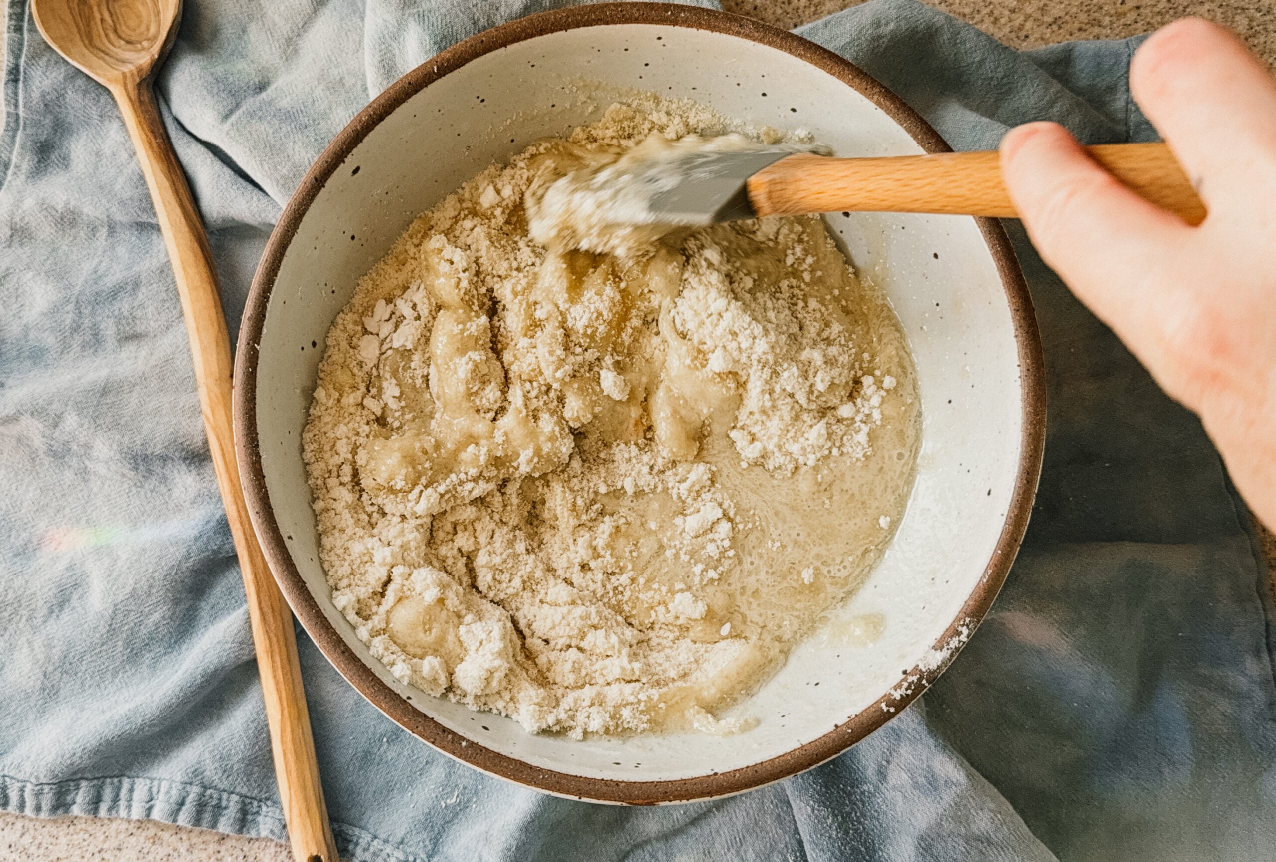 A bowl of ingredients being mixed with a spatula. A wooden spoon rests to the right of the bowl and a blue tea towel lays underneath the bowl.