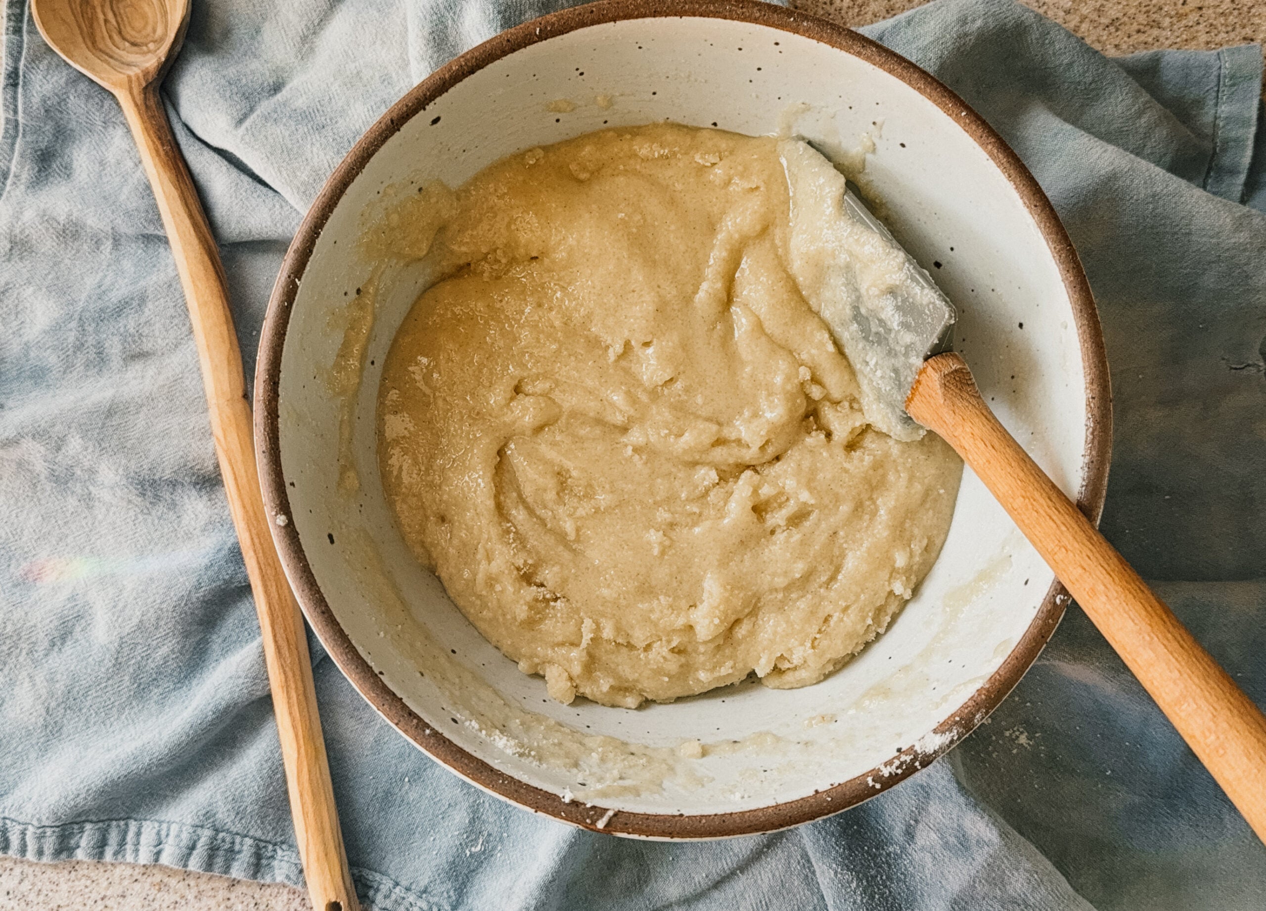 A spatula mixing cake batter. A wooden spoon rests to the left side of the bowl and a blue tea towel lays underneath.
