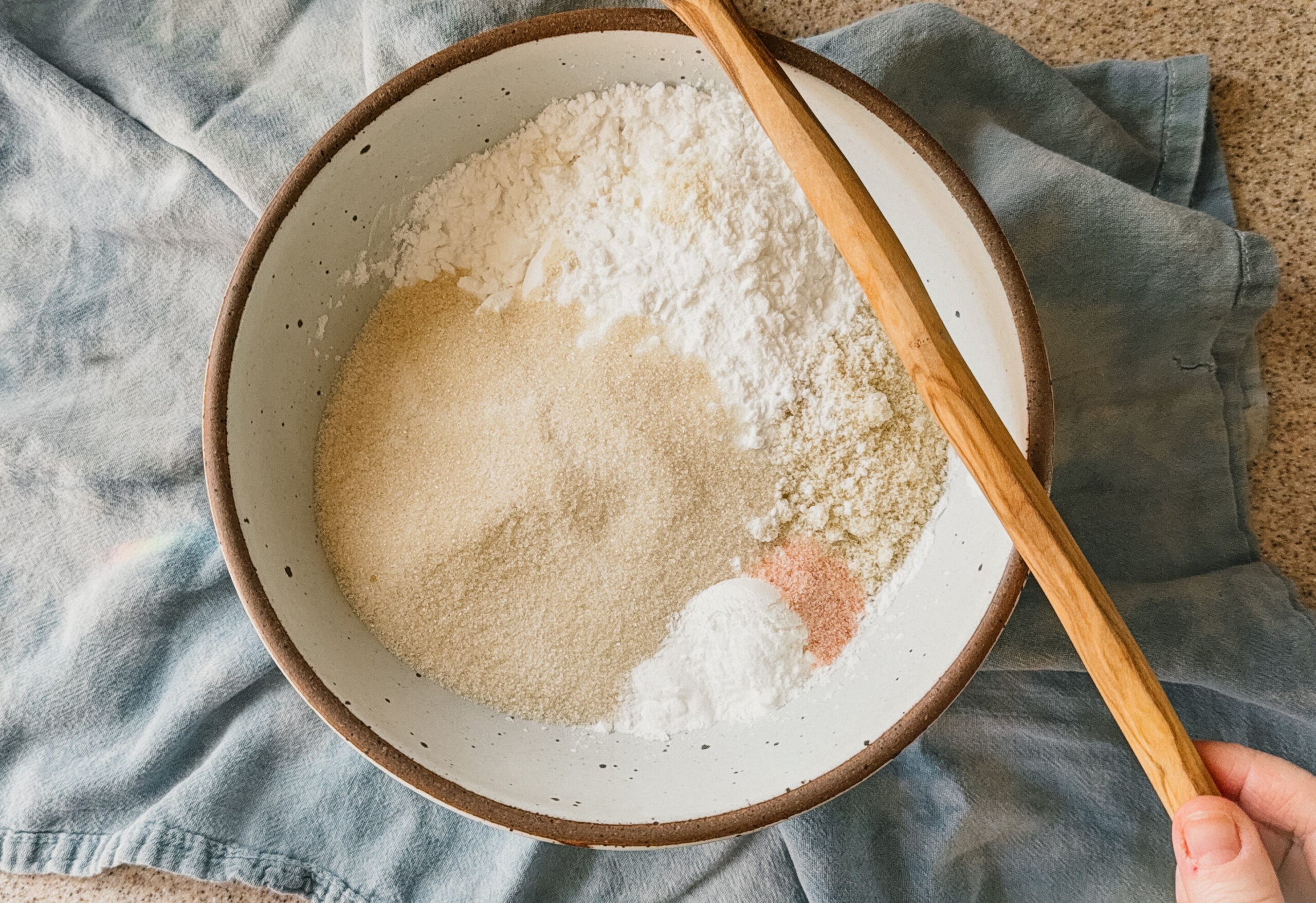 A bowl with ingredients for almond flour cake about to be mixed. A spatula rests on the right side of the bowl and a blue tea towel lays underneath.