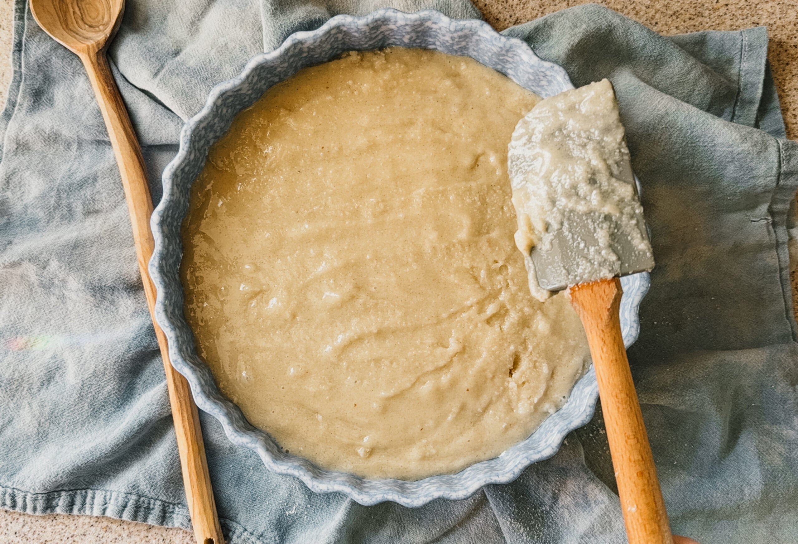 A cake pan full of batter. A spatula is to the right side of the pan, a wooden spoon rests on the left. A blue tea towel lays underneath.