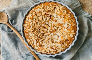 A freshly baked almond flour cake. A wooden spoon rests to the left of the cake and a blue tea towel lays underneath.