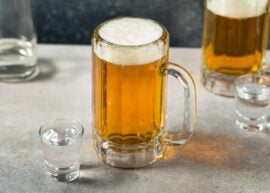 A mug of beer with foam next to a shot glass on a table.