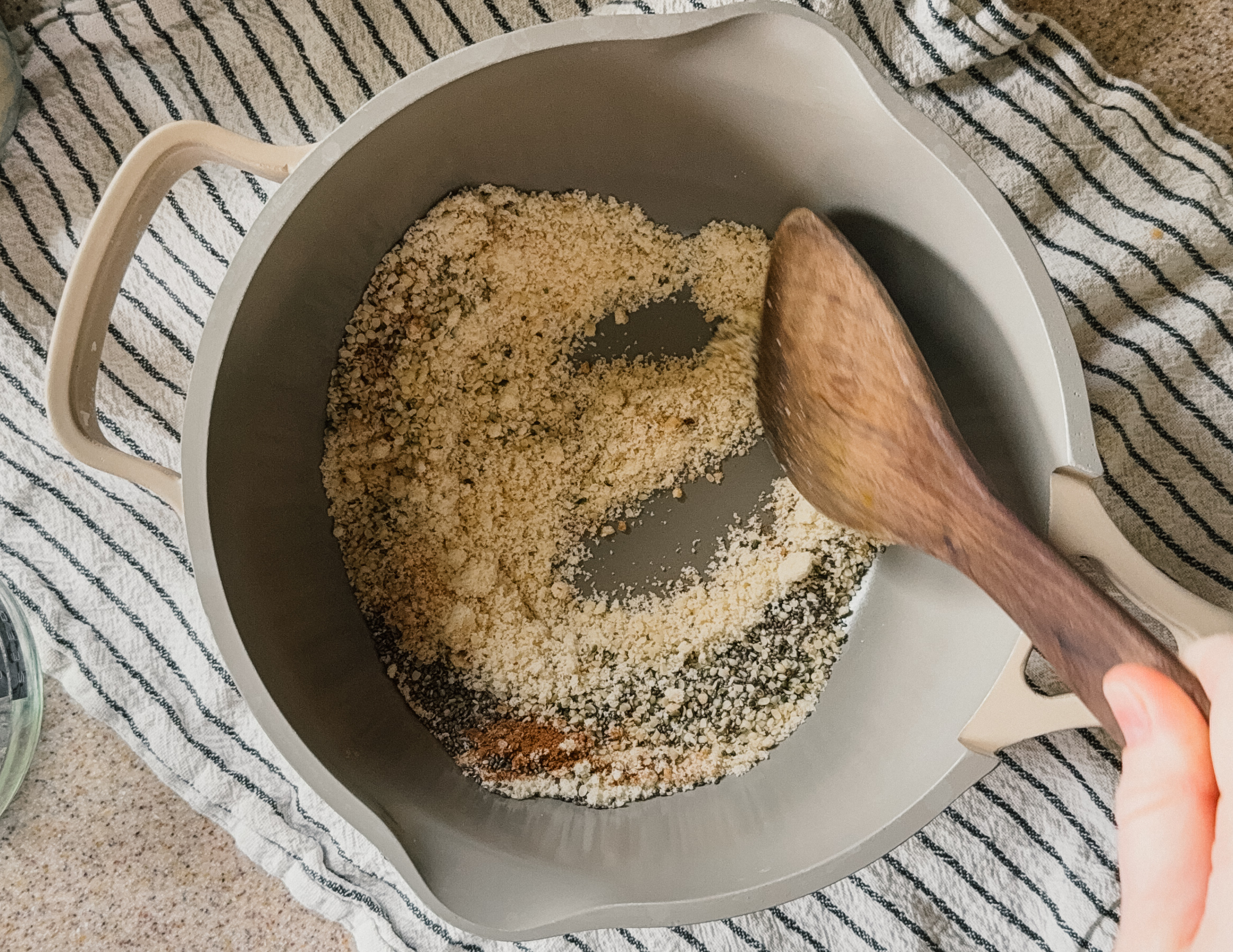 A pot of ingredients being stirred by a wooden spoon. A striped tea towel lays underneath the pot.