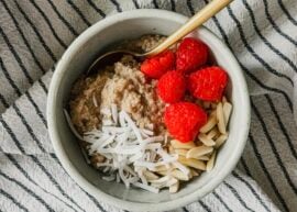 A bowl of oatmeal topped with coconut, almonds, and raspberries. A gold spoon sticks out to the right side and a striped tea towel lays underneath the bowl.