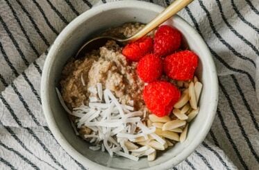 A bowl of oatmeal topped with coconut, almonds, and raspberries. A gold spoon sticks out to the right side and a striped tea towel lays underneath the bowl.