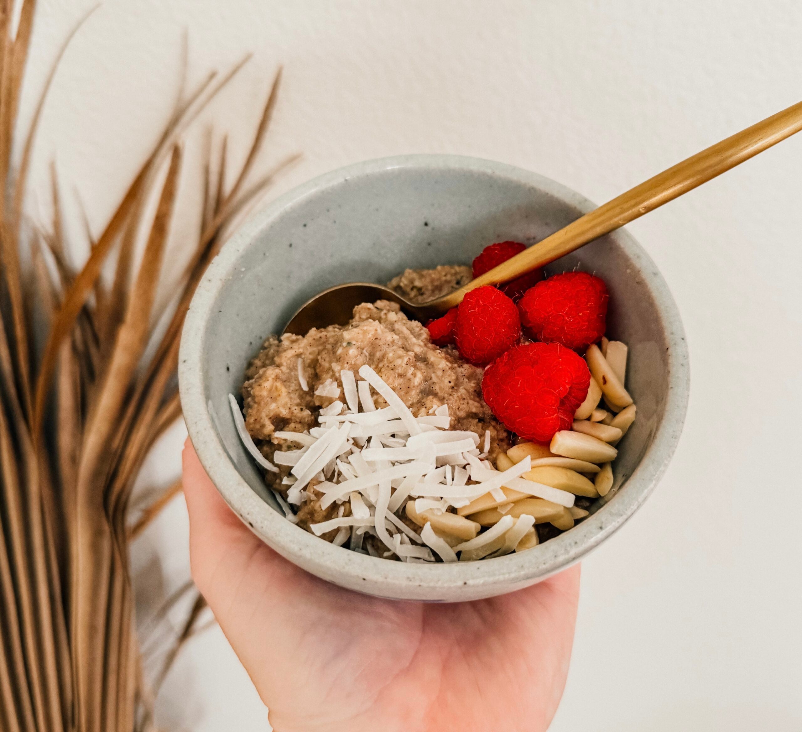 A hand holding a bowl of oatmeal topped with coconut, almonds, and raspberries. A gold spoon sticks out to the right side and a palm fronds is in the left background.