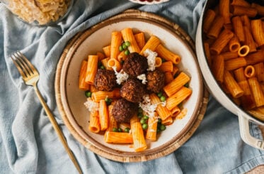 A bowl of pasta, meatballs, peas, and cheese laying on top of a tan trivet and blue tea towel. A gold fork rests to the left of the bowl and bowls of food are arranged around the top of the bowl.