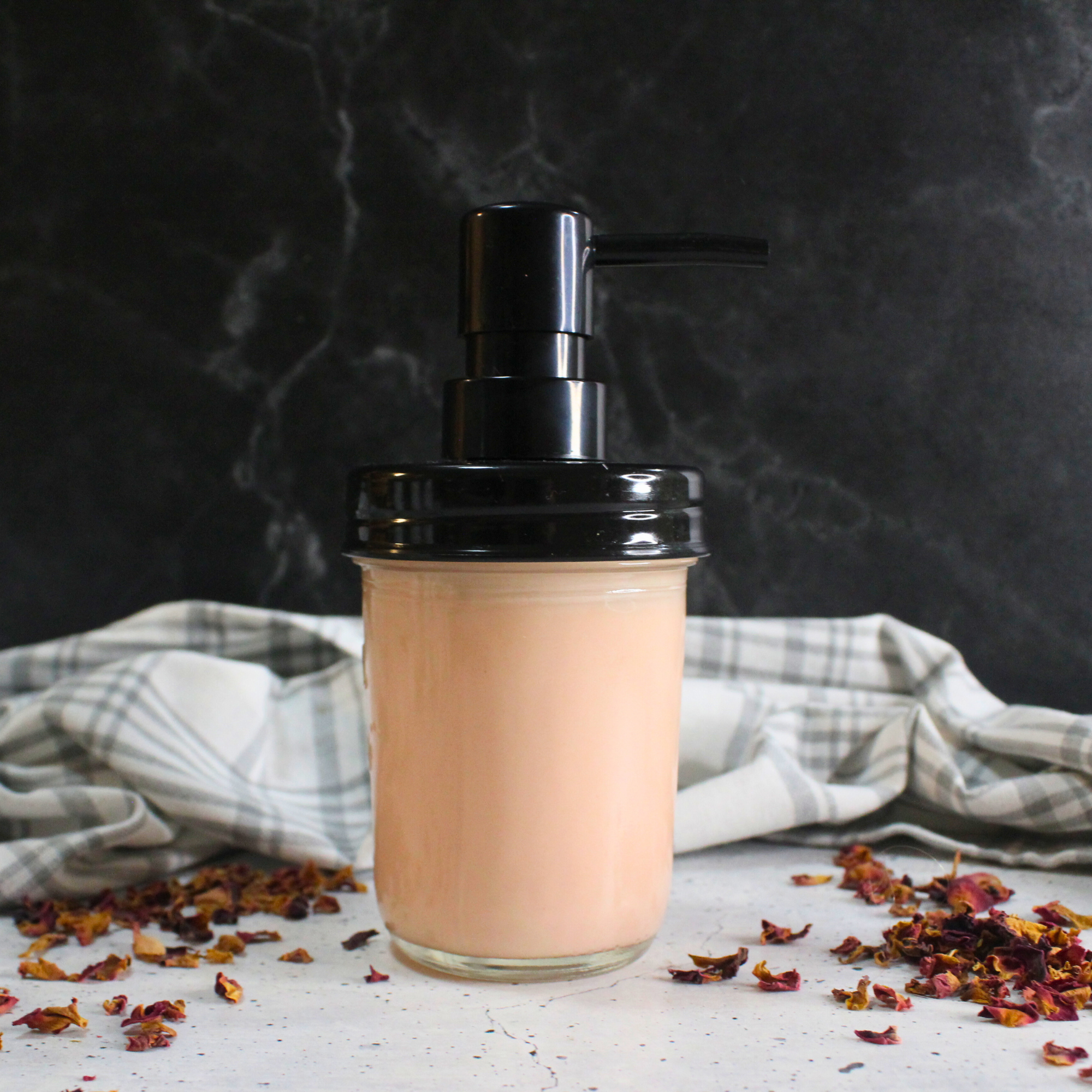 Homemade rose lotion in a mason jar with rose petals surrounding it.