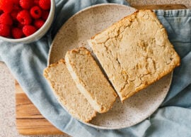 A loaf of bread on a plate with several slices. A blue tea towel lays underneath and a bowl of raspberries sits to the left of the bread.