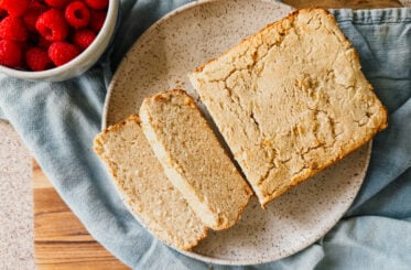 A loaf of bread on a plate with several slices. A blue tea towel lays underneath and a bowl of raspberries sits to the left of the bread.