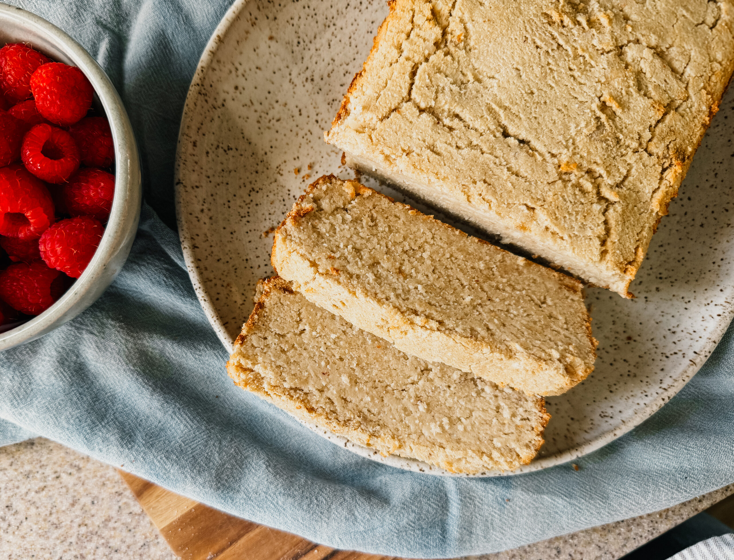A loaf of bread on a plate with several slices. A blue tea towel lays underneath and a bowl of raspberries sits to the left of the bread.