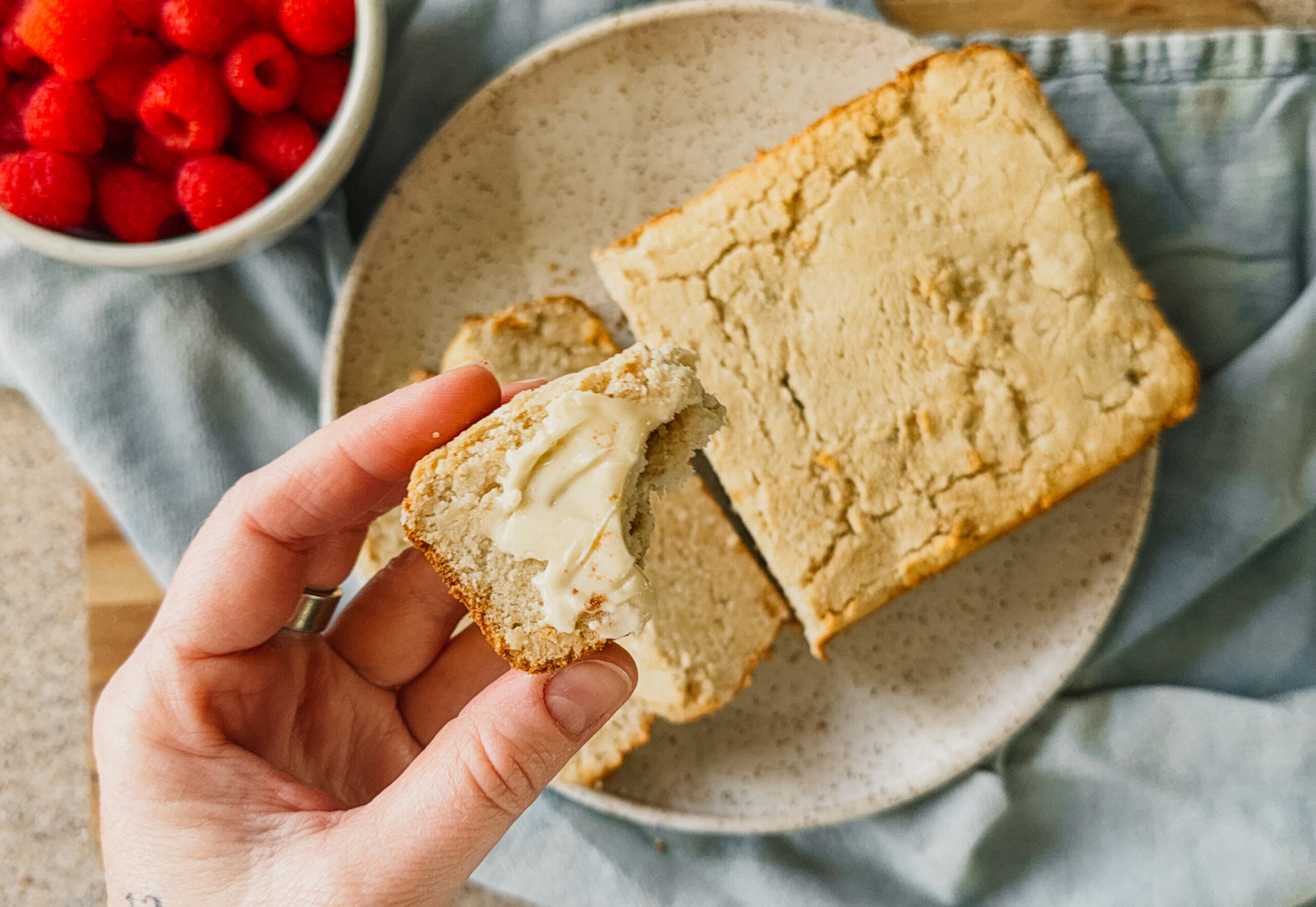 A hand holding a bitten piece of buttered bread above the remaining loaf of bread on a plate. A blue tea towel lays underneath and a bowl of raspberries sits to the left of the bread.