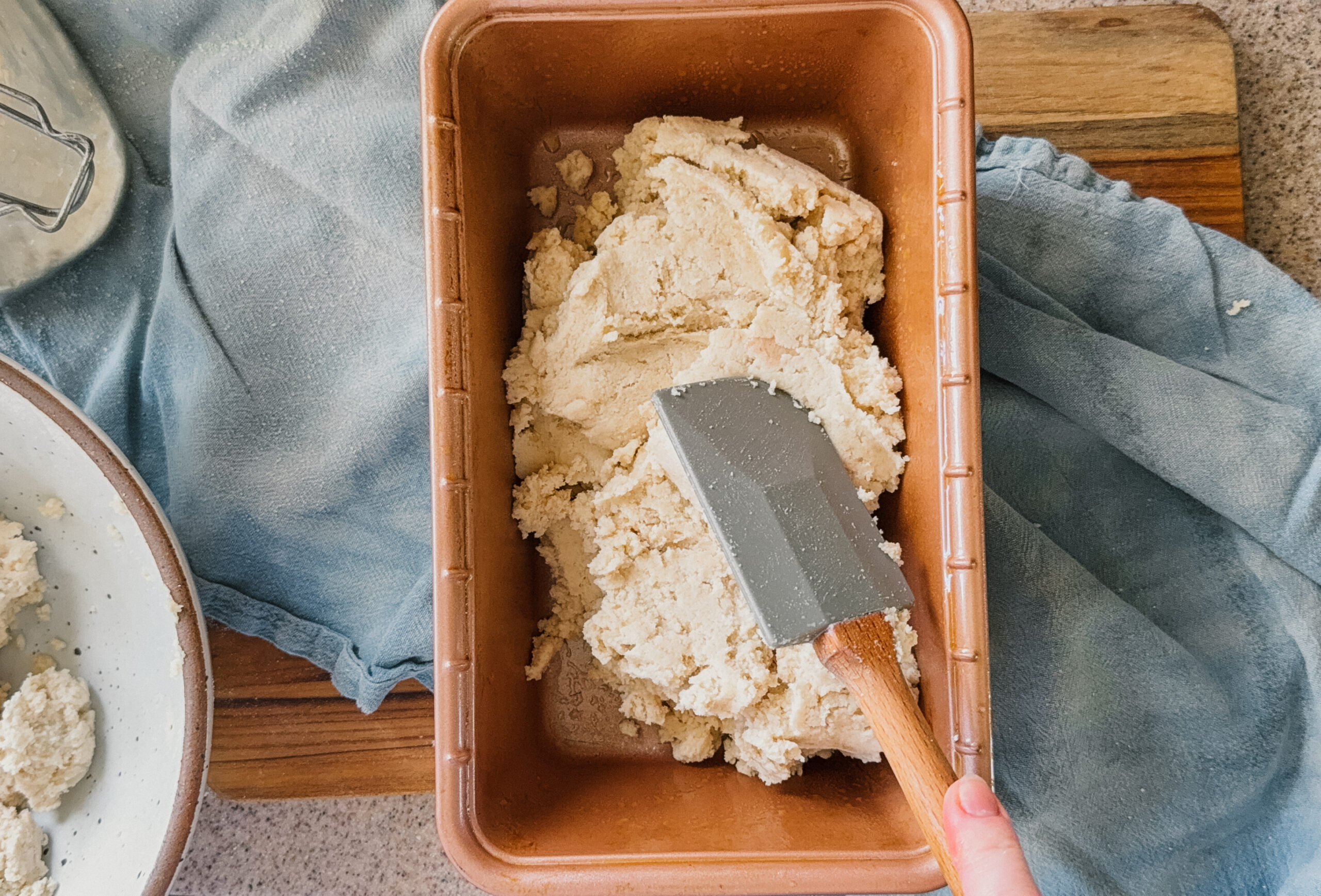 Bread dough being scooped into a bread pan with a spatula. A blue tea towel and wooden board are underneath the pan.