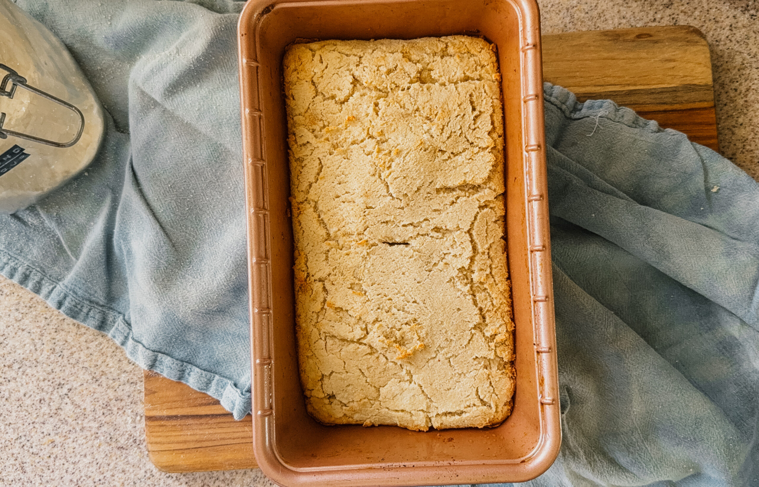 Freshly baked bread in a bread pan sitting on top of a blue tea towel and wooden board.