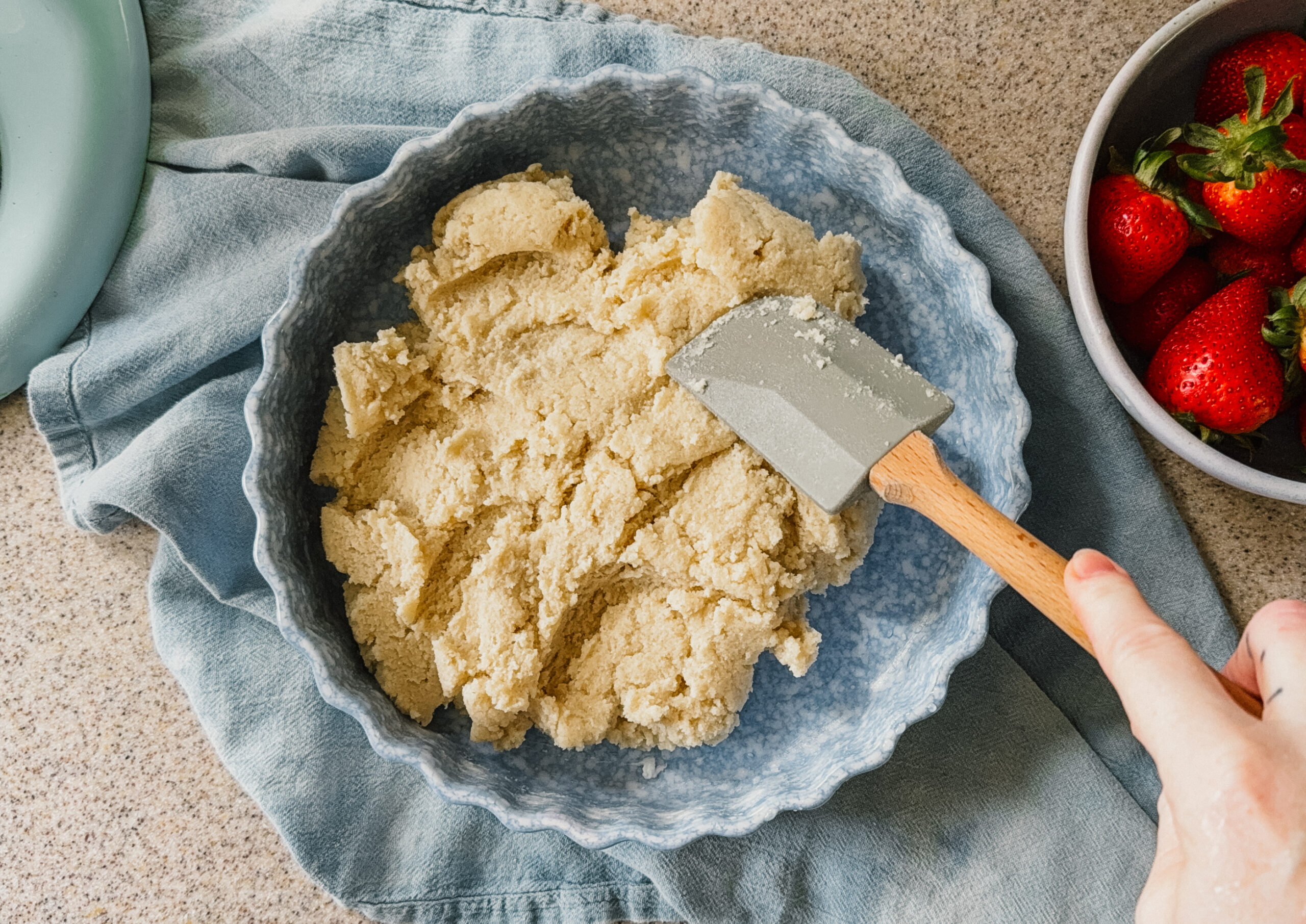 Cake batter being pressed into a cake dish with a spatula. A blue tea towel lies underneath the dish and a bowl of strawberries is in the upper right corner.