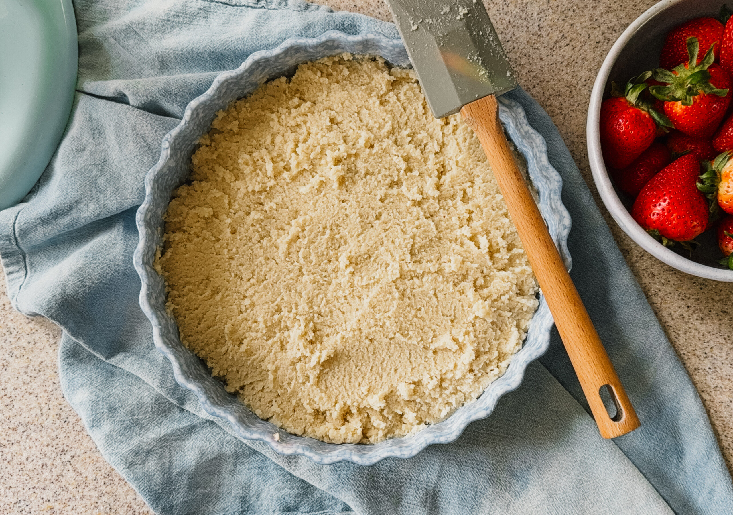 Cake batter in a cake dish about to be baked. A blue tea towel lies underneath the dish and a spatula rests on the right hand side. A bowl of strawberries is in the upper right corner.