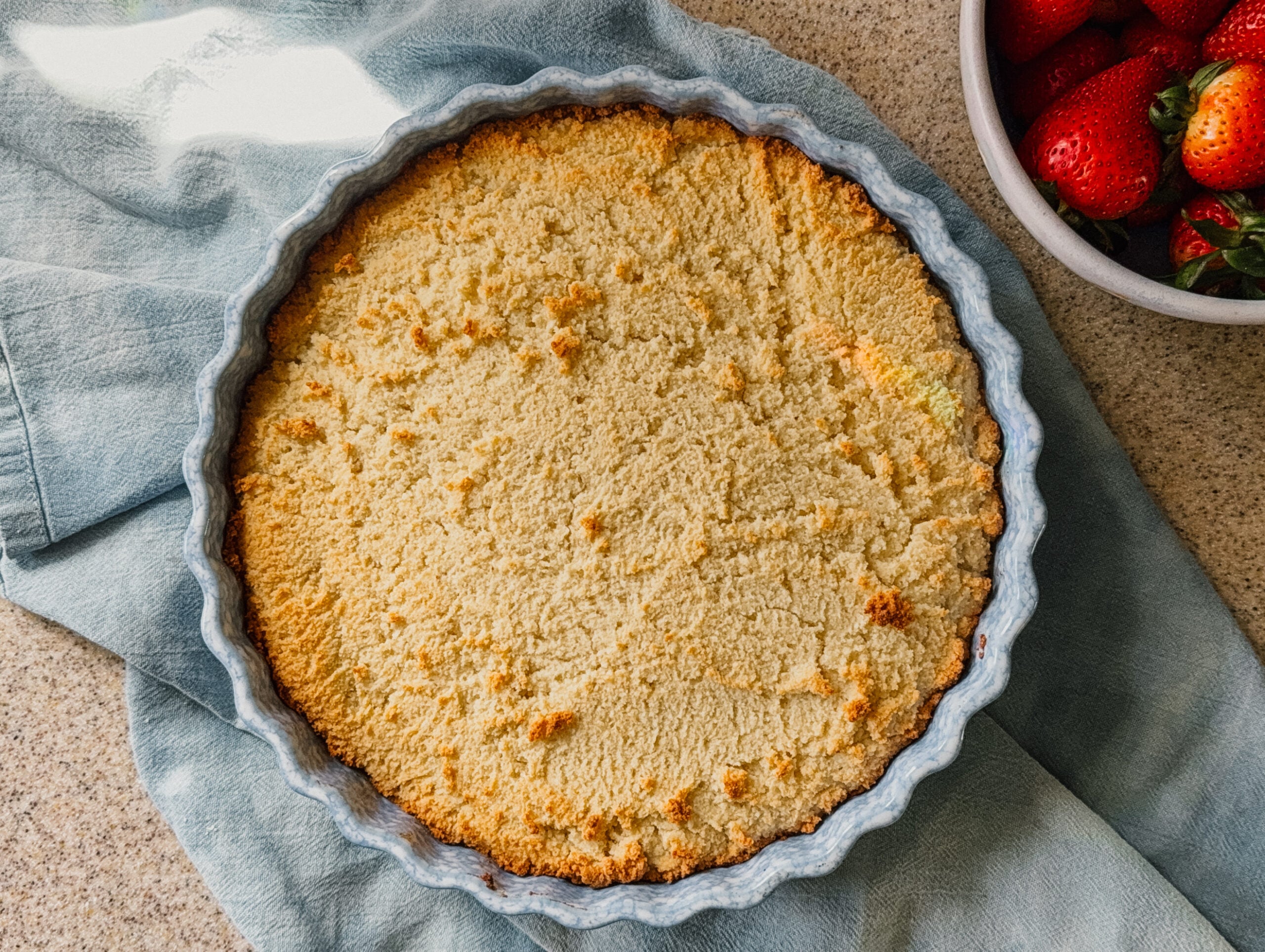 A freshly baked cake in a dish on top of a blue tea towel. A bowl of strawberries is in the top right.