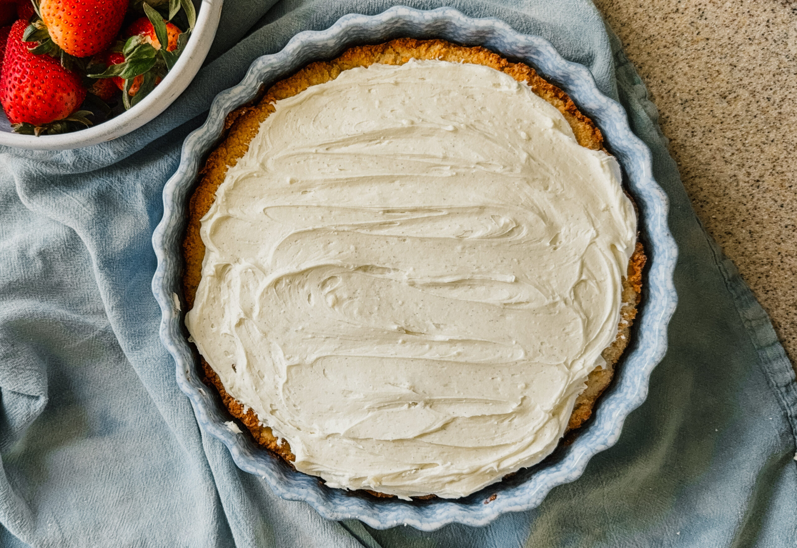 A freshly frosted cake in a cake dish. A blue tea towel lies underneath the dish and a bowl of strawberries is in the upper left corner.