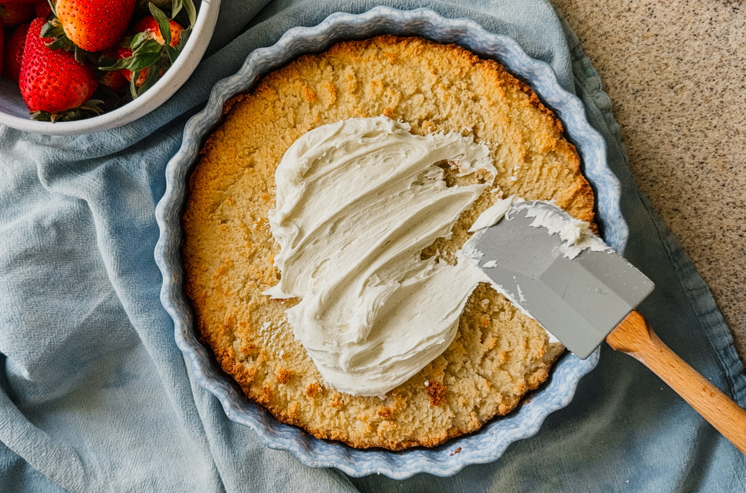 A spatula frosting a cake in a cake dish. A blue tea towel lies underneath the dish and a bowl of strawberries is in the upper left corner.