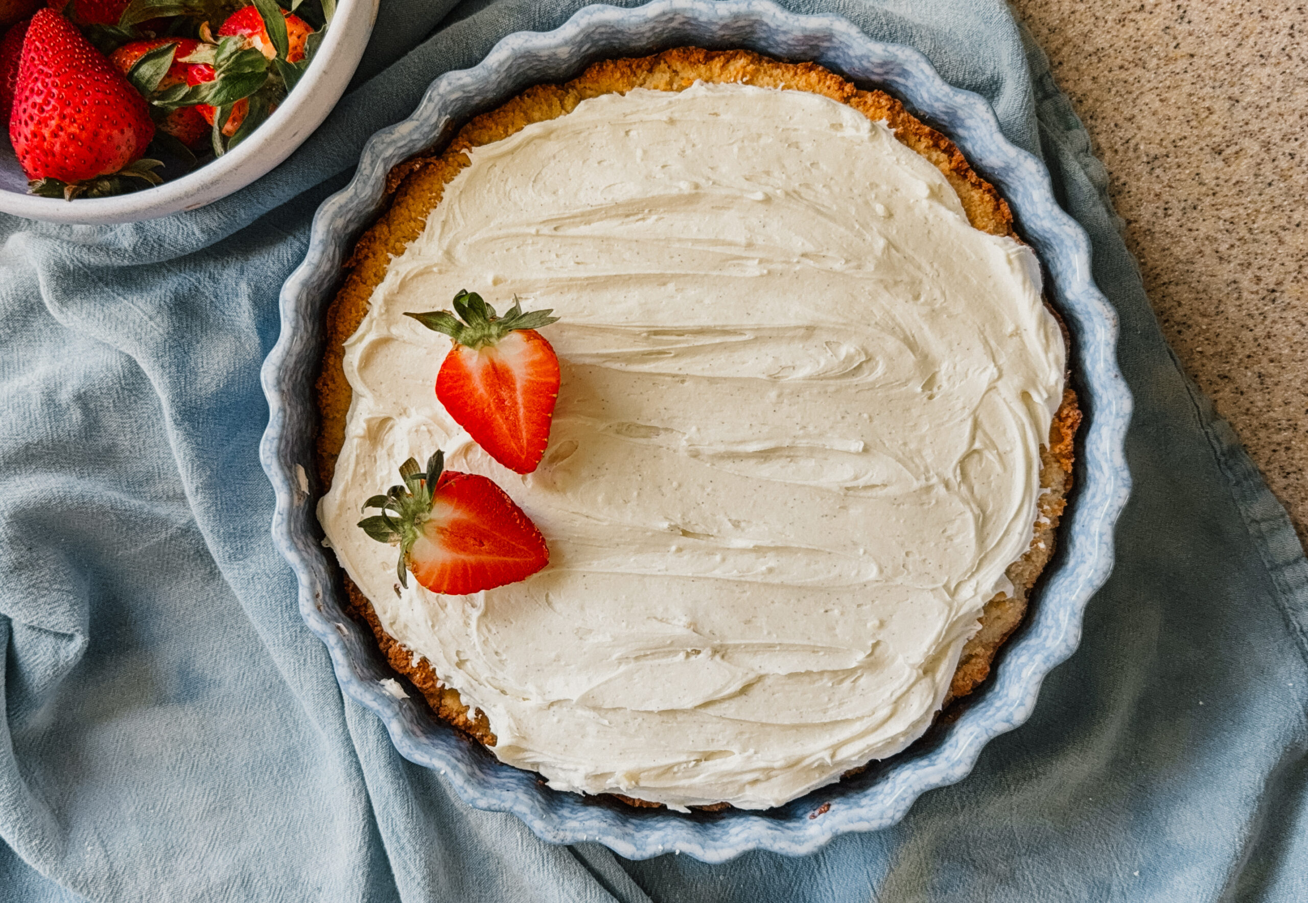 A frosted cake in a cake dish with two strawberries on its left side. A blue tea towel lies underneath the dish and a bowl of strawberries is in the upper left corner.