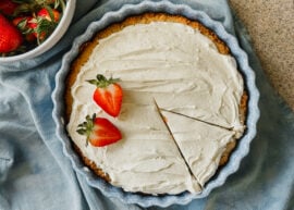 A frosted cake in a cake dish with two strawberries on its left side. A slice has been cut in the bottom right of the cake and a blue tea towel lies underneath the dish. A bowl of strawberries is in the upper left corner.