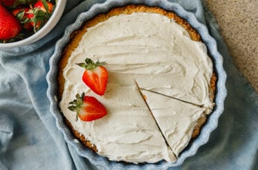 A frosted cake in a cake dish with two strawberries on its left side. A slice has been cut in the bottom right of the cake and a blue tea towel lies underneath the dish. A bowl of strawberries is in the upper left corner.