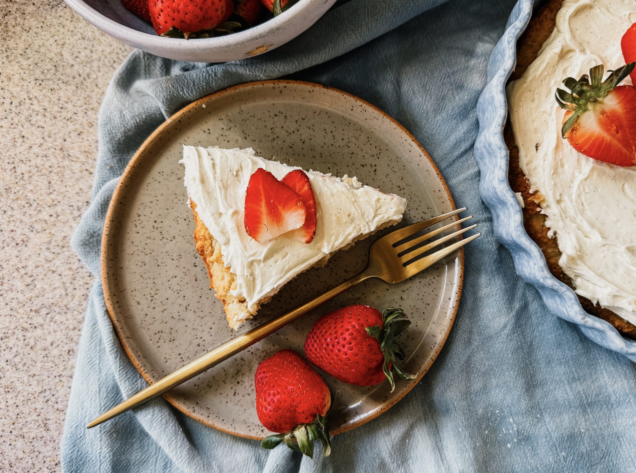 A slice of cake on a plate. Strawberries are on top of the slice and on the lower portion of the plate, along with a gold fork. 