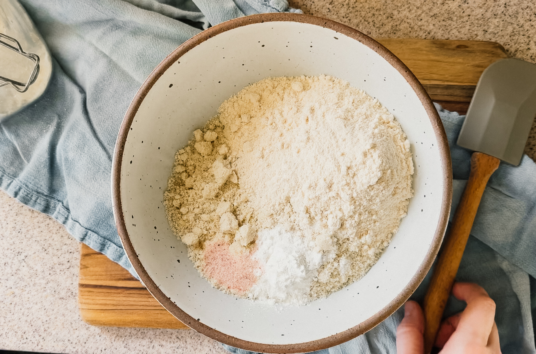 A bowl of dry ingredients sitting on top of a blue tea towel and wooden board. A hand reaches for a spatula on the right hand side.