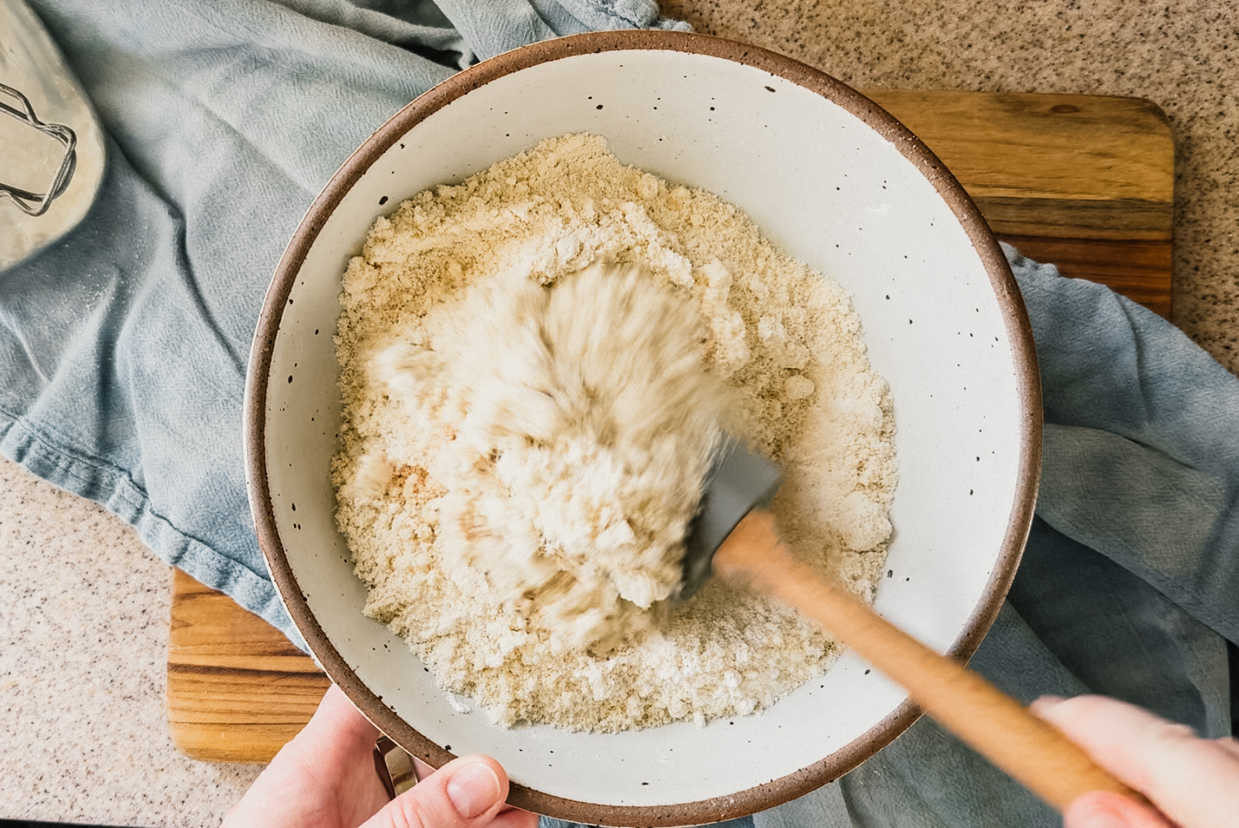A bowl of dry ingredients being mixed. A blue tea towel and wooden board sit underneath the bowl. A hand holds the bottom of the bowl and another hand holds a spatula in the bottom right.
