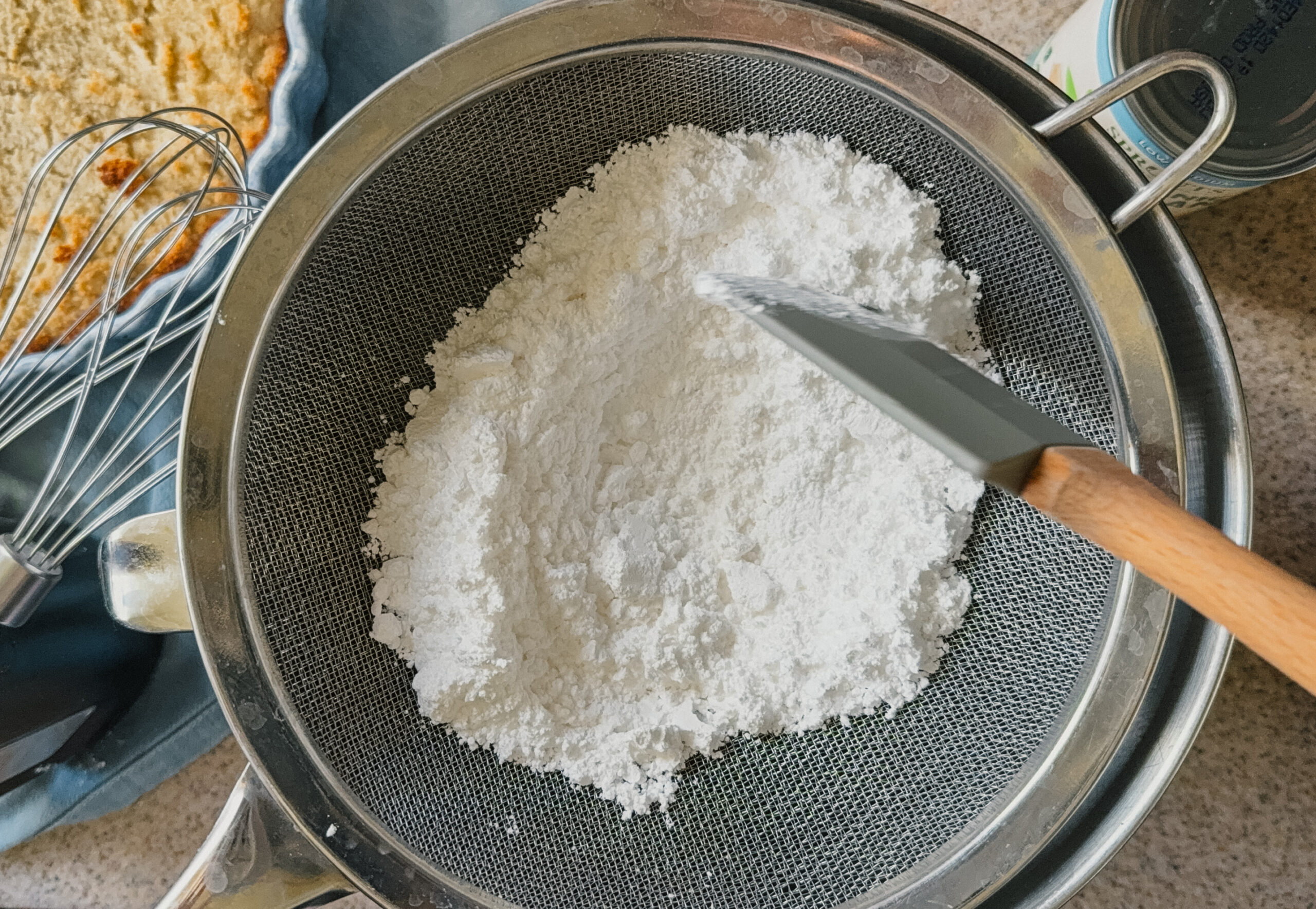 Powdered sugar being sifted through a sifter into a bowl with a spatula.