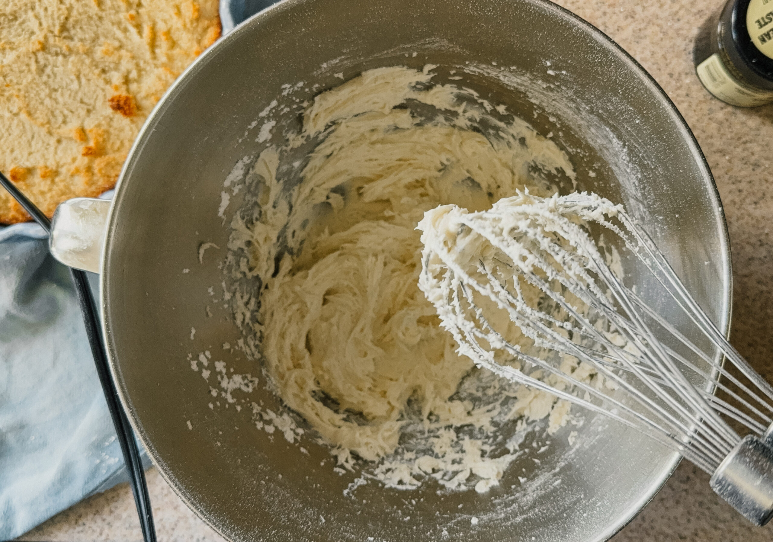 A whisk covered in frosting hovering above a bowl with the remaining frosting.