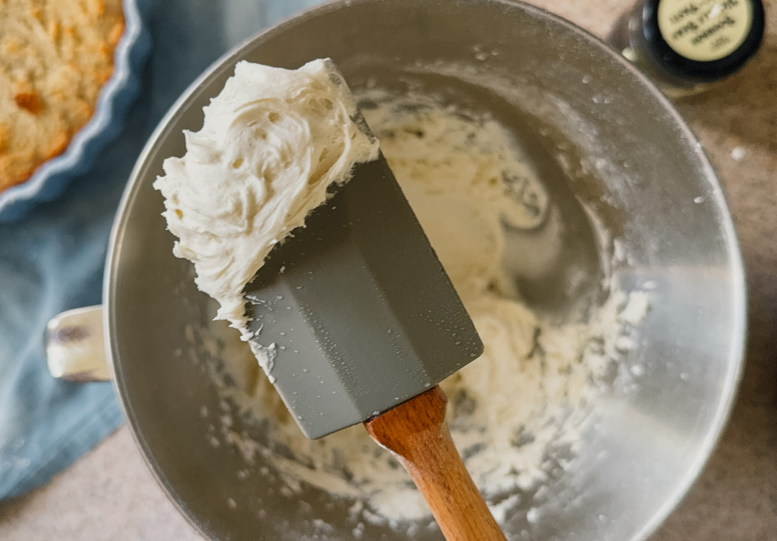 A spatula with a dollop of frosting hovering above a mixer bowl with the remaining frosting.