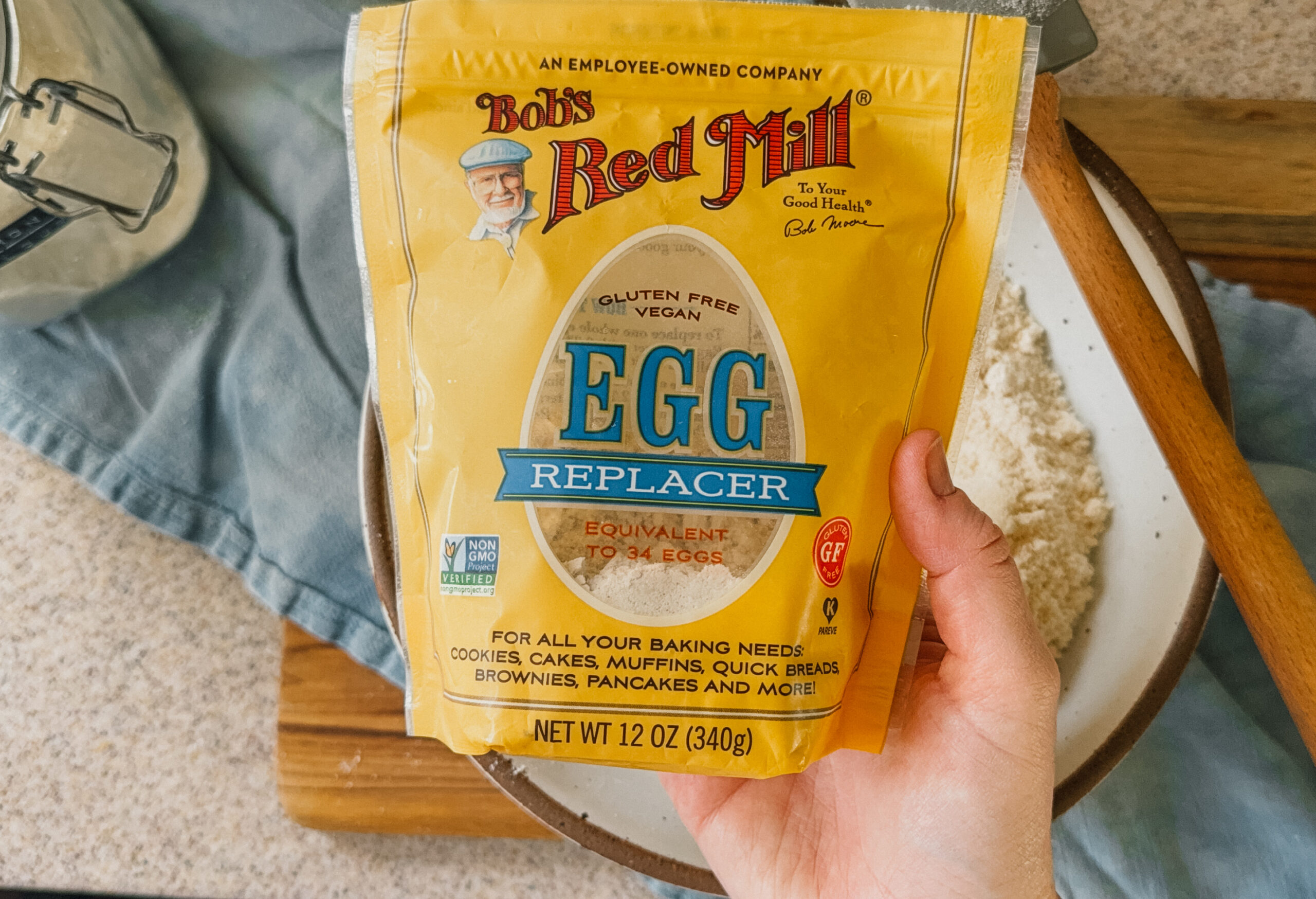 A hand holding a bag of egg replacer powder. A bowl, spatula, blue tea towel, and wooden board are in the background.