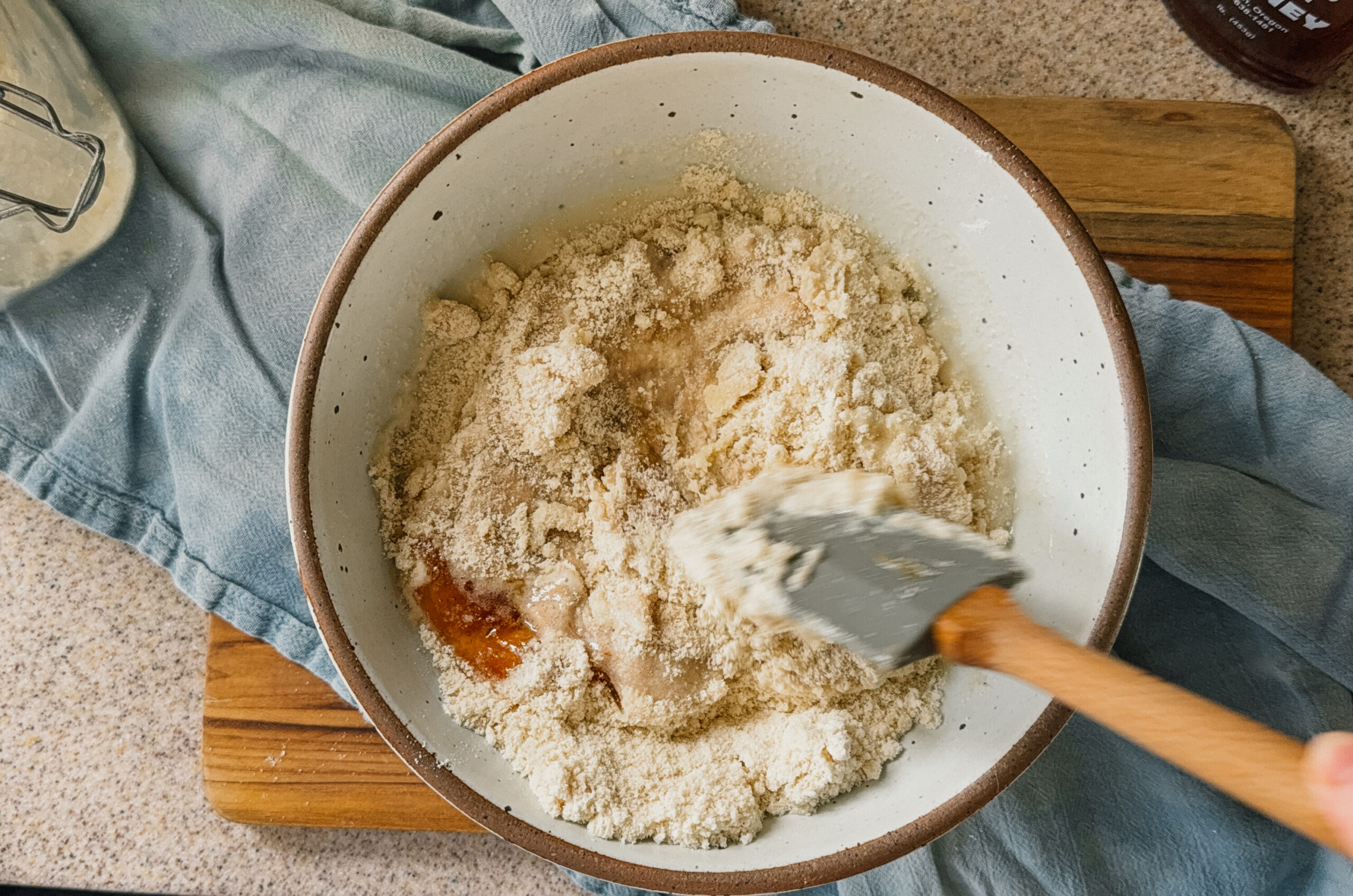A bowl of ingredients being mixed with a spatula. A blue tea towel and wooden board sit underneath the bowl. A hand holds the bottom of the bowl and another hand holds a spatula in the bottom right.