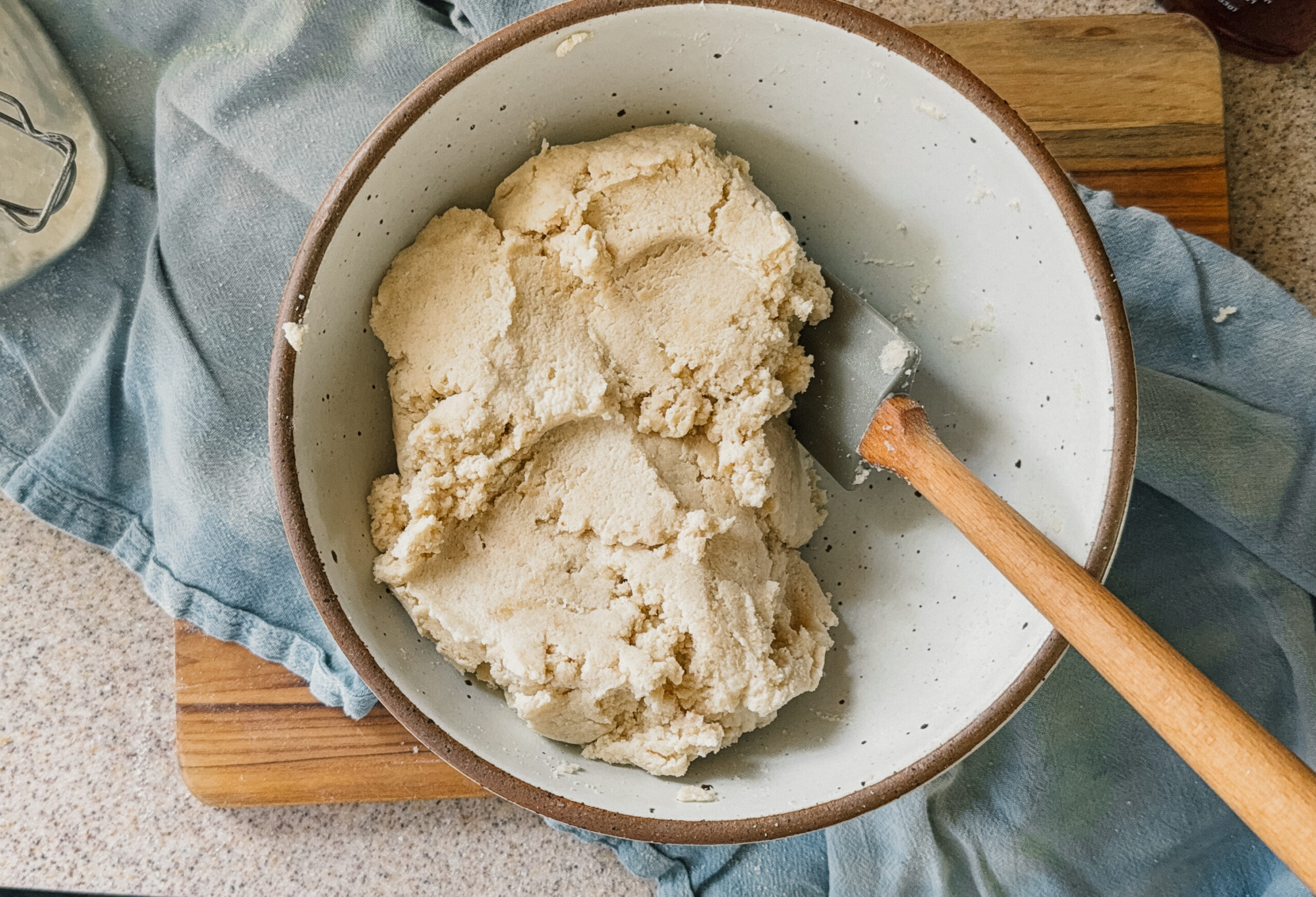 A bowl of dough with a spatula sticking out of the right side. A blue tea towel and wooden board are underneath the bowl.