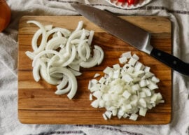 Two piles of onion pieces, one sliced and the other diced, on a wooden cutting board. A large knife lies in the upper right corner.