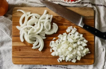 Two piles of onion pieces, one sliced and the other diced, on a wooden cutting board. A large knife lies in the upper right corner.