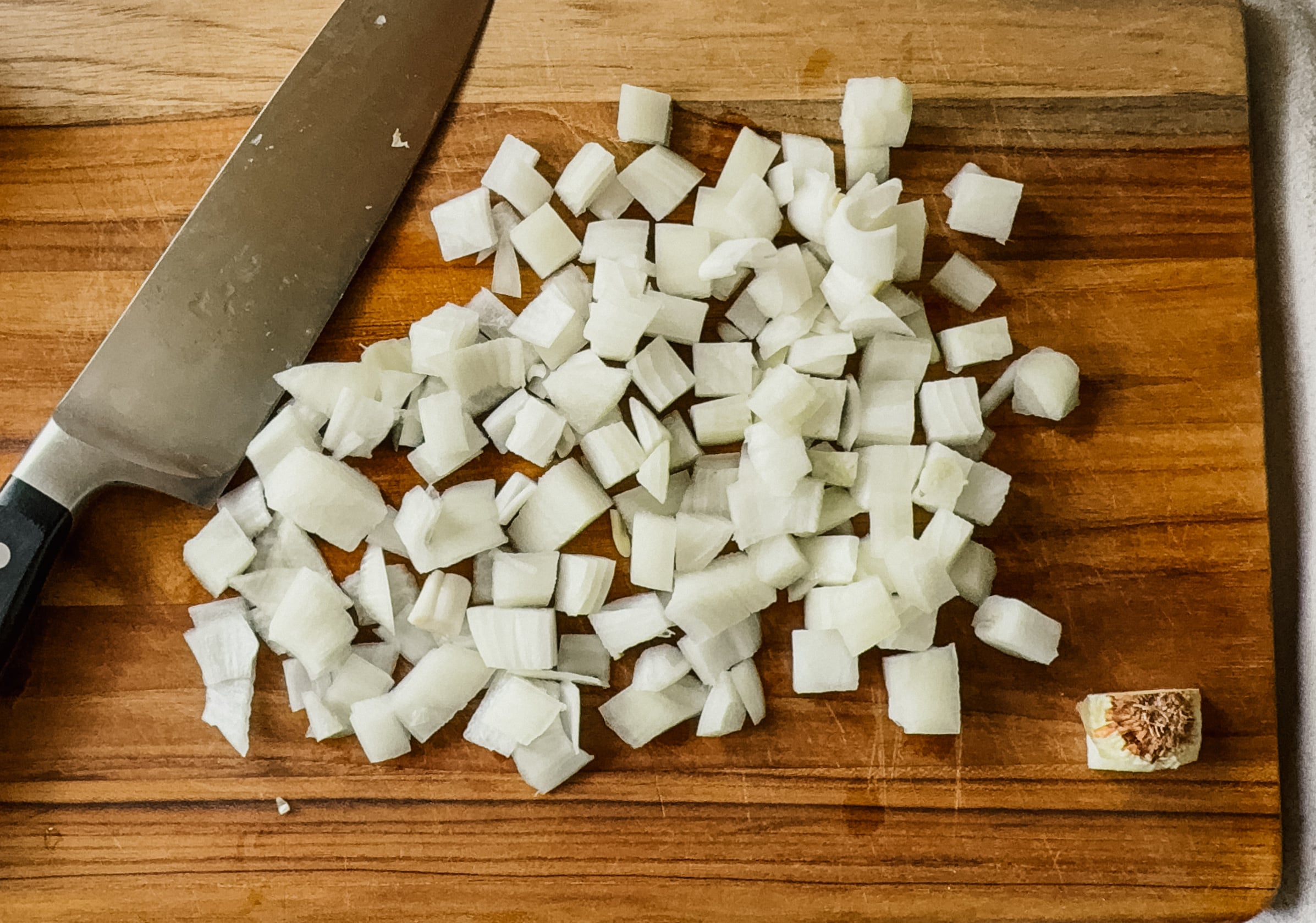 A pile of diced onion on a wooden cutting board. A large knife lies to the left hand side.