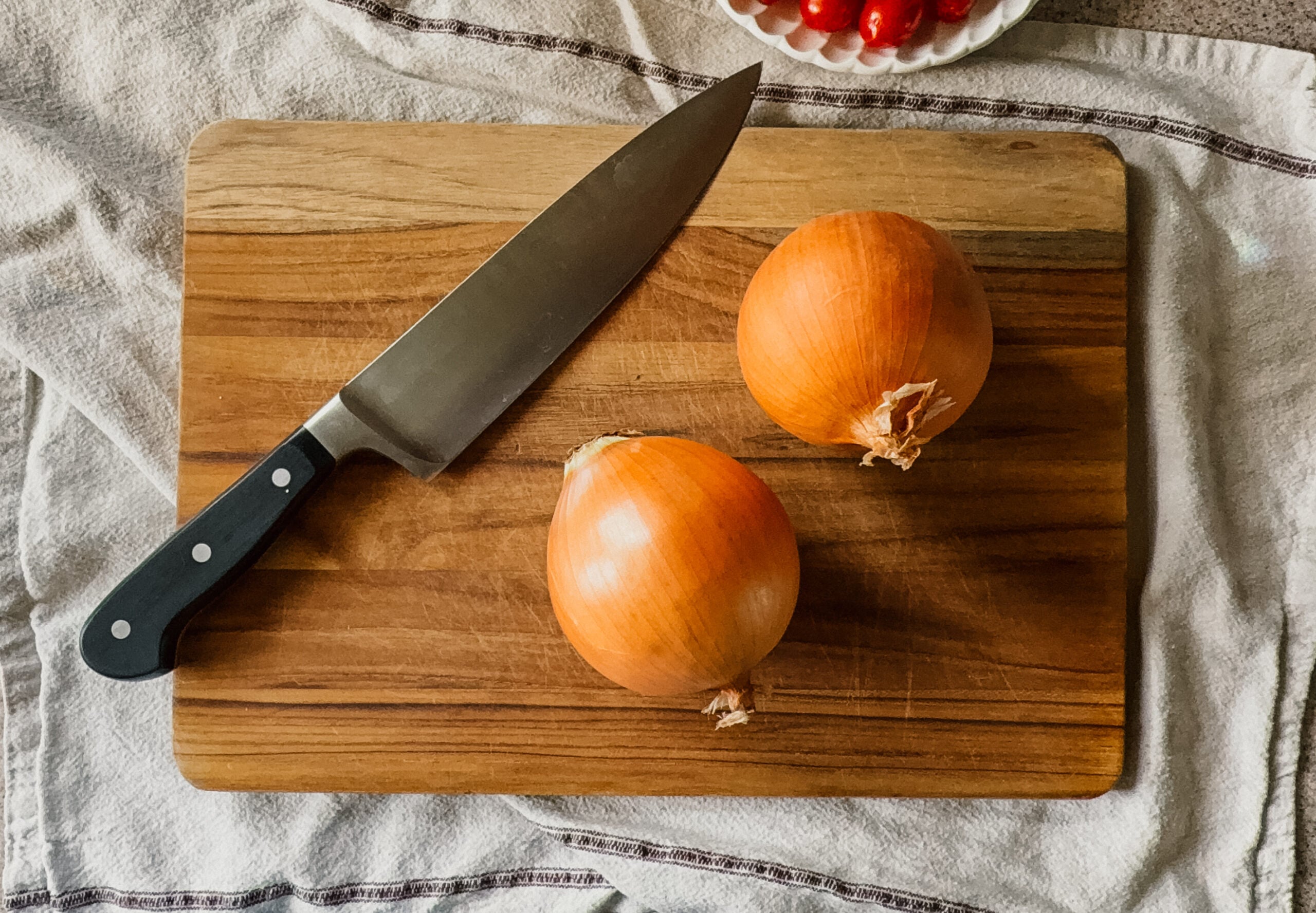 Two yellow onions and a large knife on top of a wooden cutting board. A tan tea towel lays underneath the board.