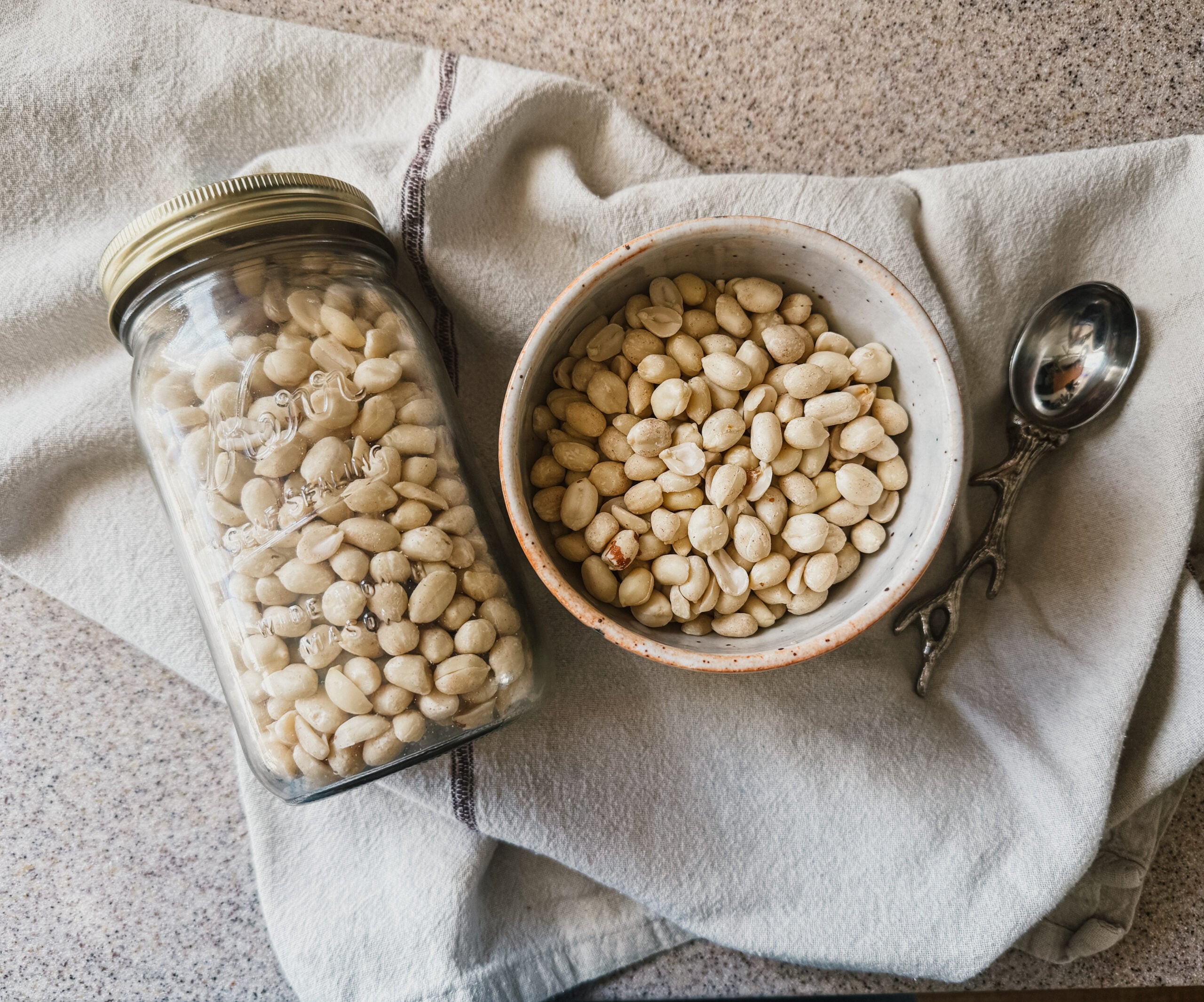 A jar of peanuts next to a bowl of peanuts. Both lay on top of a brown tea towel.