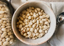 A bowl of peanuts on top of a brown tea towel. A jar of peanuts is to the left of the bowl.