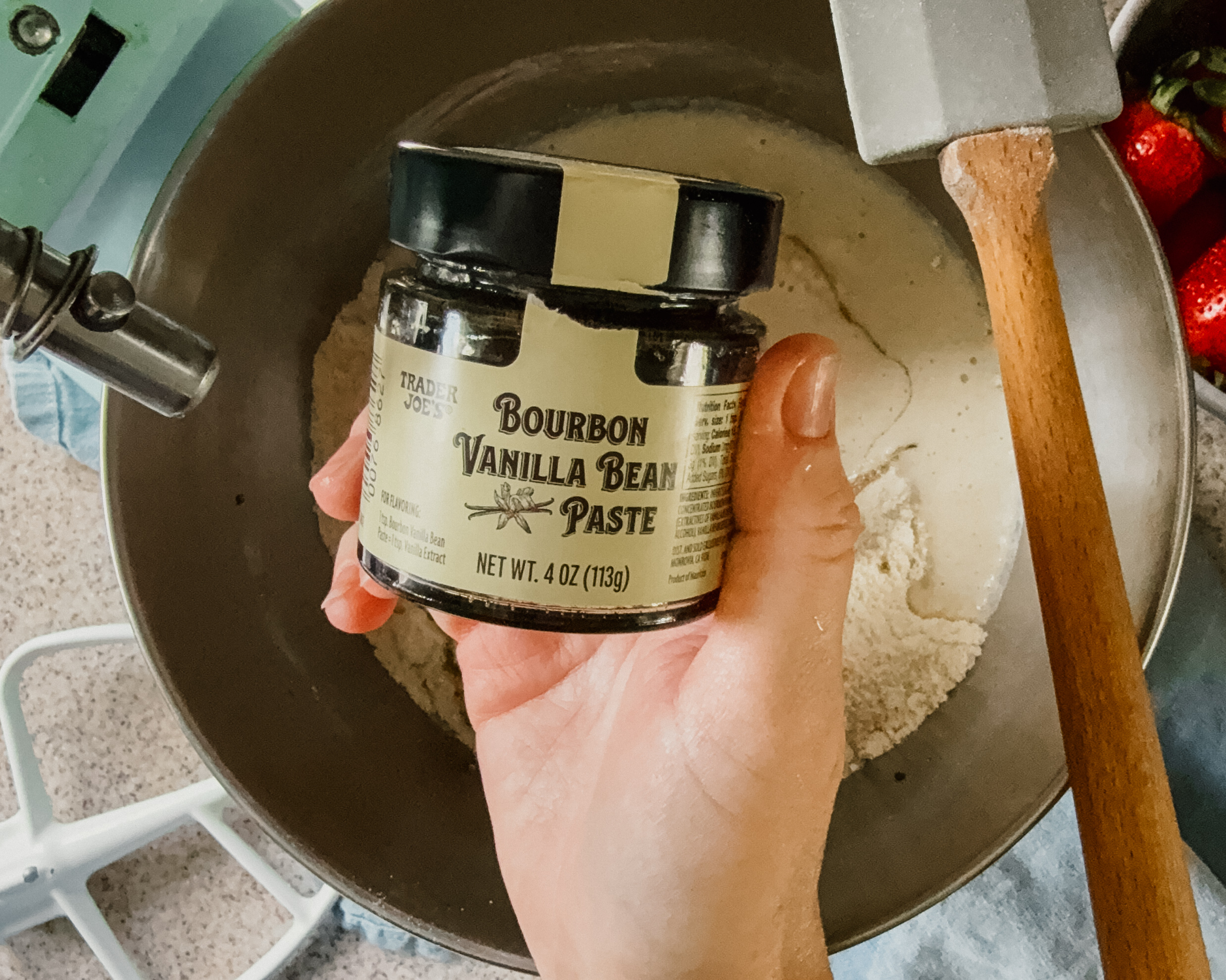 A hand holding a small jar of vanilla bean extract above a mixer bowl with ingredients. A spatula rests on the right side of the bowl.