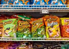 Multiple bags of Cheetos snacks on store shelves, including Puffs, Flamin' Hot, and Mexican Street Corn flavors.