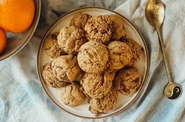A plate of cookies on top of a blue tea towel. A bowl of oranges is in the top left corner.
