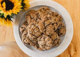 A plate of freshly baked cookies on a white plate. A vase of sunflowers is in the upper left corner.